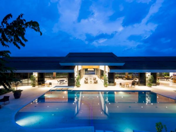 The image shows a luxurious modern building with a large, illuminated swimming pool in front, set against a twilight sky.