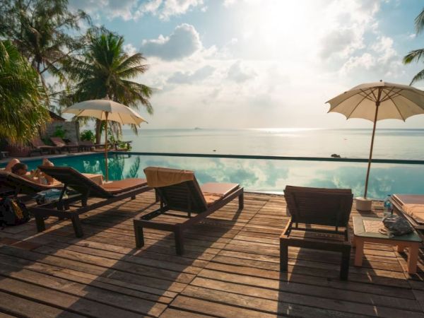 A serene seaside scene with lounge chairs, umbrellas on a wooden deck, palm trees, and a calm ocean beneath a partly cloudy sky ending the sentence.