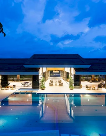 A luxurious modern building with a large illuminated swimming pool and seating area at dusk, reflecting the serene blue sky.