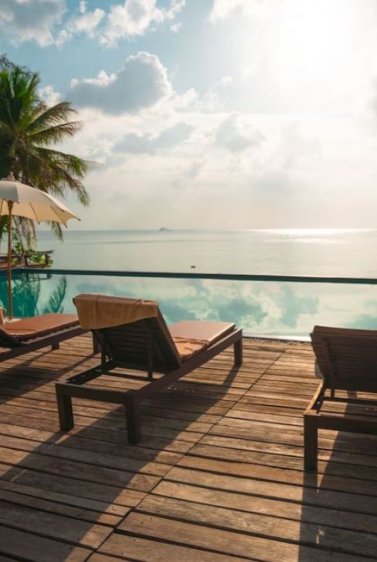 The image shows a serene poolside scene with lounge chairs, umbrellas, and a view of the ocean under a partly cloudy sky, surrounded by palm trees.