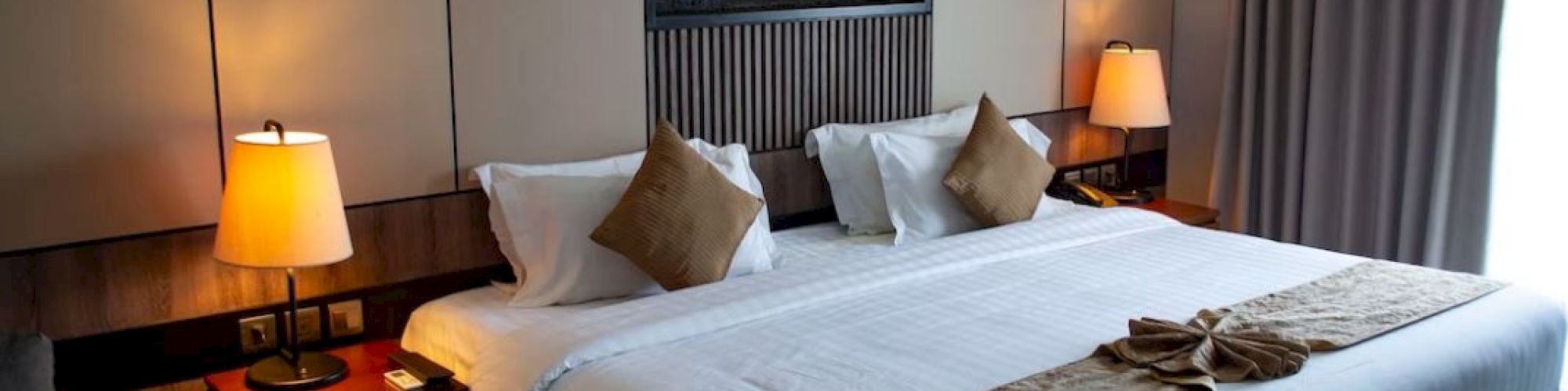 A hotel room with a large bed, bedside tables with lamps, and an abstract wall art piece above the bed. There's a window with curtains closed.