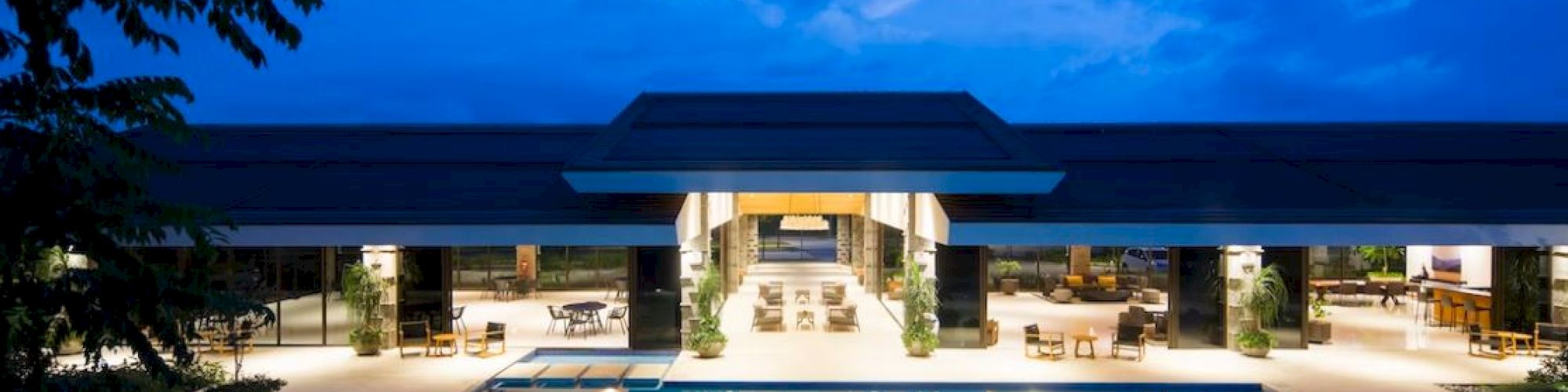 Elegant building with an illuminated pool in the foreground, surrounded by seating areas and trees, under a blue evening sky.
