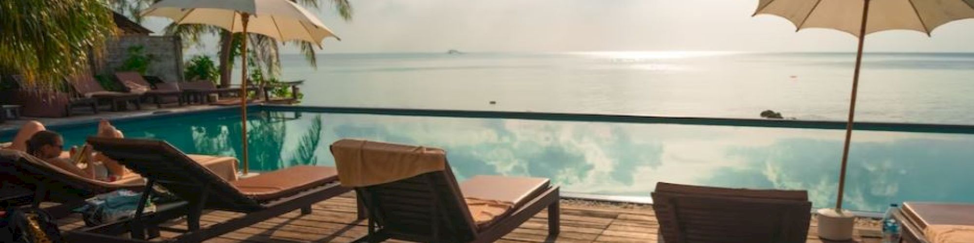 A serene beachfront scene with lounge chairs, umbrellas, and an infinity pool overlooking the sea, surrounded by palm trees under a partly cloudy sky.