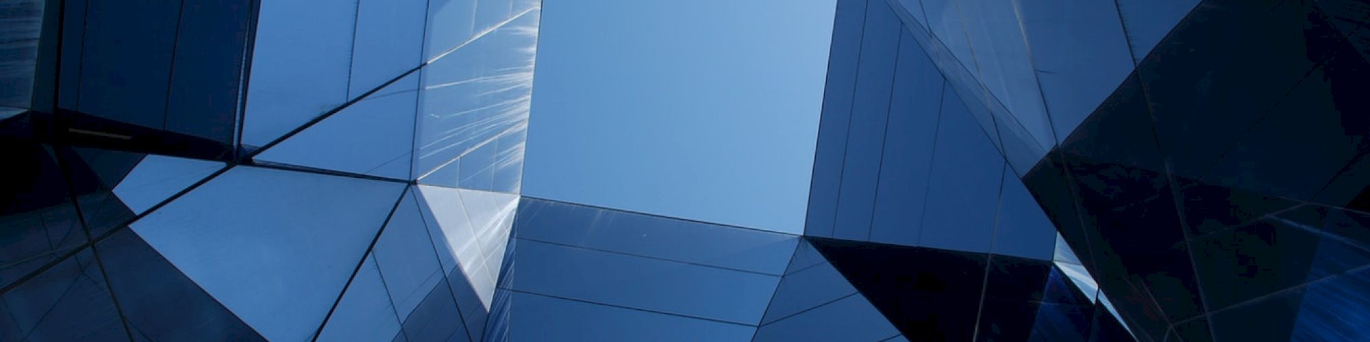 The image shows an upward view of modern glass buildings with a geometric design, creating a pattern that frames a clear blue sky at the center.