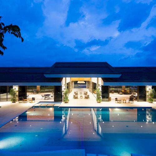 The image shows a luxurious modern building with an illuminated swimming pool at dusk, surrounded by seating and lounge areas.
