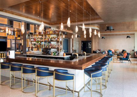 A modern bar with pendant lights, blue bar stools, and a stocked liquor wall. There's additional seating in the background and a sleek interior design.