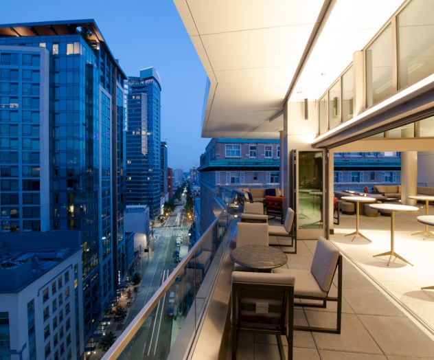 A cityscape at dusk showing a modern high-rise balcony with seating, a view of adjacent buildings, and a street below lit by streetlights.