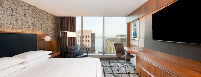 A modern hotel room with a large bed, desk, chair, flat-screen TV, and a view of city buildings through a floor-to-ceiling window.
