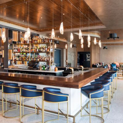 This image shows a modern bar with a wooden counter, blue and gold barstools, hanging lights, and a well-stocked back bar.