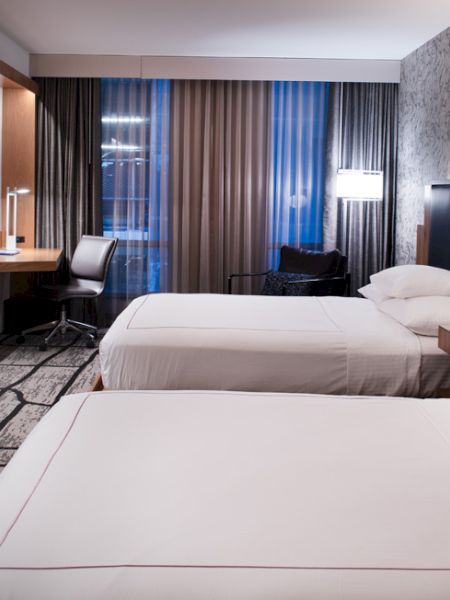 A modern hotel room with two neatly made beds, a wall-mounted TV, a desk with a chair, bedside lamps, and a large window with curtains.