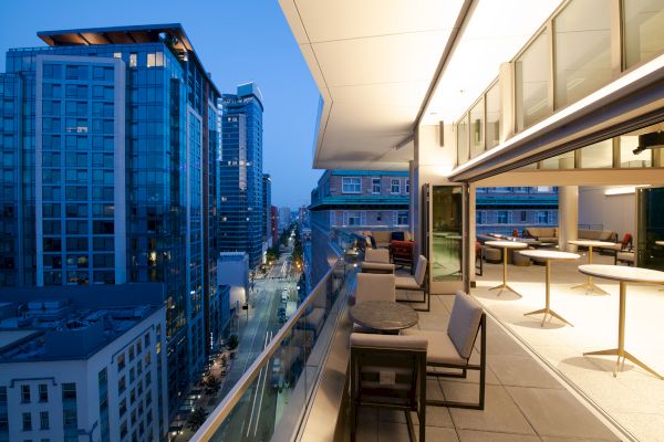 The image features a modern urban balcony with seating, overlooking a cityscape with tall buildings illuminated in the evening.