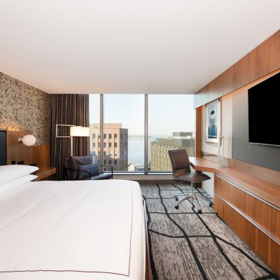 A modern hotel room features a large bed, a wall-mounted TV, a desk, a chair, and a window with a city view.