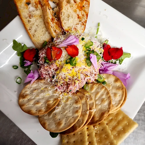 A plate with toasted bread, round and square crackers, cheese, shredded meat, strawberries, purple flowers, and greens in the center.