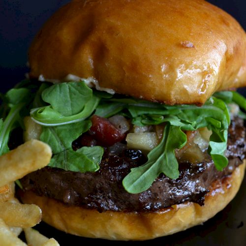A juicy burger topped with greens, cheese, and bacon, beside crispy fries on a dark background.