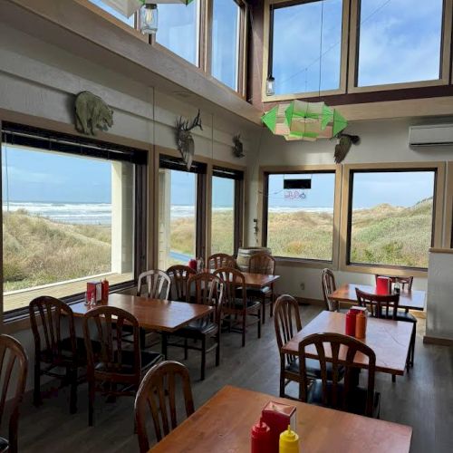 A seaside caf&eacute; with wooden tables and chairs, condiments on tables, large windows with ocean views, and green ceiling d&eacute;cor.