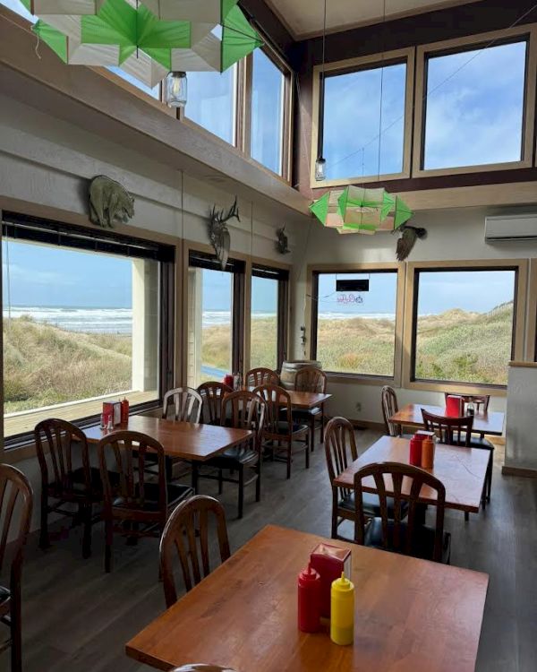 A seaside caf&eacute; with wooden tables and chairs, condiments on tables, large windows with ocean views, and green ceiling d&eacute;cor.