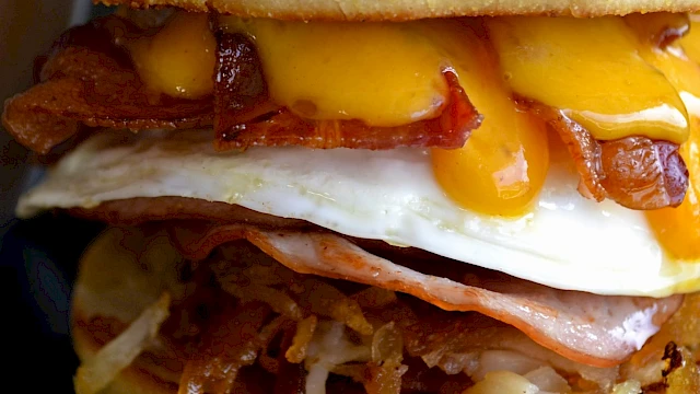 A close-up bacon cheeseburger with melted cheese, fried onions, crispy bacon, a fried egg, and caramelized onions in a toasted bun.