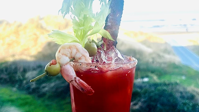 A tall red cocktail with ice and a fancy herb garnish, a slice and fruit skewers, against a bright outdoor coastal backdrop. Top it at 140 characters, always ending the sentence.