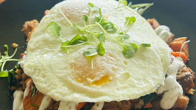 A plated stack with a fried egg on top, crispy potato and veggie base, white drizzle sauce, and green microgreens garnish on a black plate.