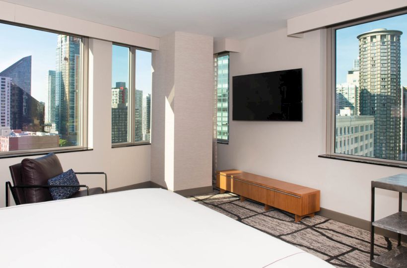 A bright, modern room with large windows showcasing a cityscape, a wall-mounted TV, a chair, a bed, and minimalistic furniture ending the sentence.