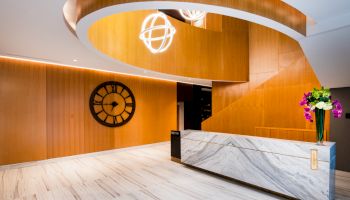 A modern lobby with a marble reception desk, large black wall clock, wooden accents, spiral staircase, and decorative lighting fixtures.