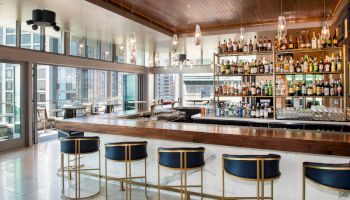 The image shows a modern bar with high stools, a well-stocked shelf of liquor bottles, and large windows offering a city view.