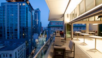 An outdoor terrace with seating overlooks a cityscape with tall buildings and a street below, captured during the evening.