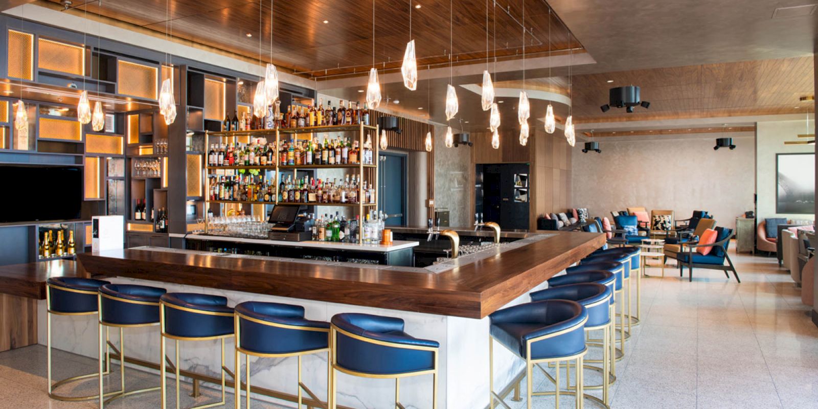 This image shows a modern bar with hanging lights, a fully stocked back bar, high-backed bar stools, and a lounge seating area in the background.