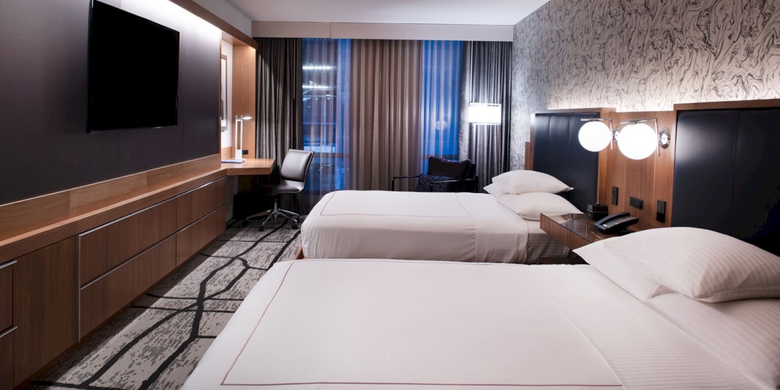 A modern hotel room with two beds, a wall-mounted TV, a desk with a chair, and stylish decor, featuring large windows with curtains.