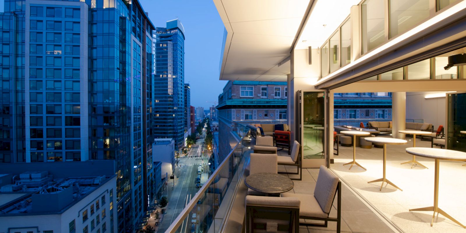 A rooftop terrace with seating and tables, overlooking a city street lined with modern buildings at dusk. The space is warmly lit.