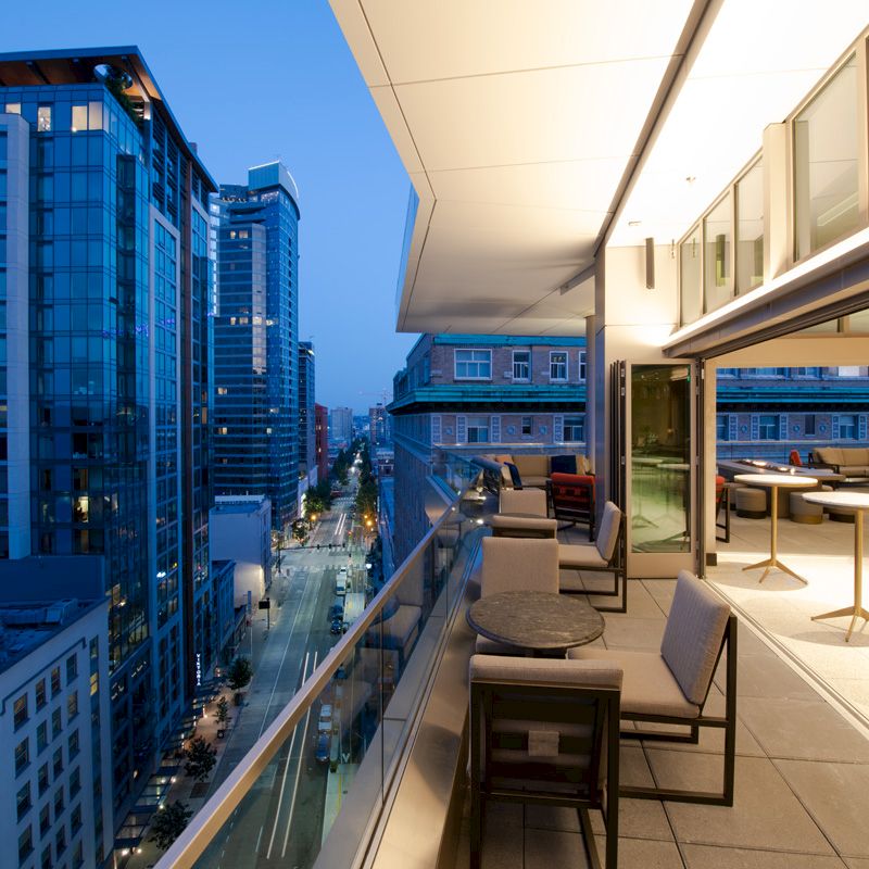 This image shows a modern balcony with furniture and cityscape view, lined with tall buildings illuminated in the early evening light.