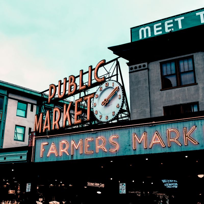 Image of a public market with neon signs reading "PUBLIC MARKET" and "FARMERS MARKET." Visible buildings and a partially hidden clock are also present.