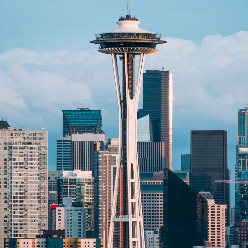 The image features the Space Needle against a backdrop of various buildings and skyscrapers in a cityscape, with a cloudy sky in the background.