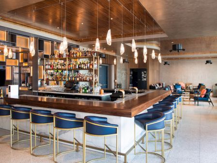 This image shows a modern bar with blue and gold bar stools, hanging light fixtures, and a well-stocked bar counter in a spacious area.