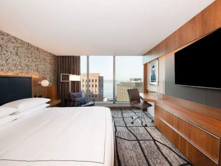 A modern hotel room with a large bed, a desk with a chair, a wall-mounted TV, and a window offering a view of city buildings and water.