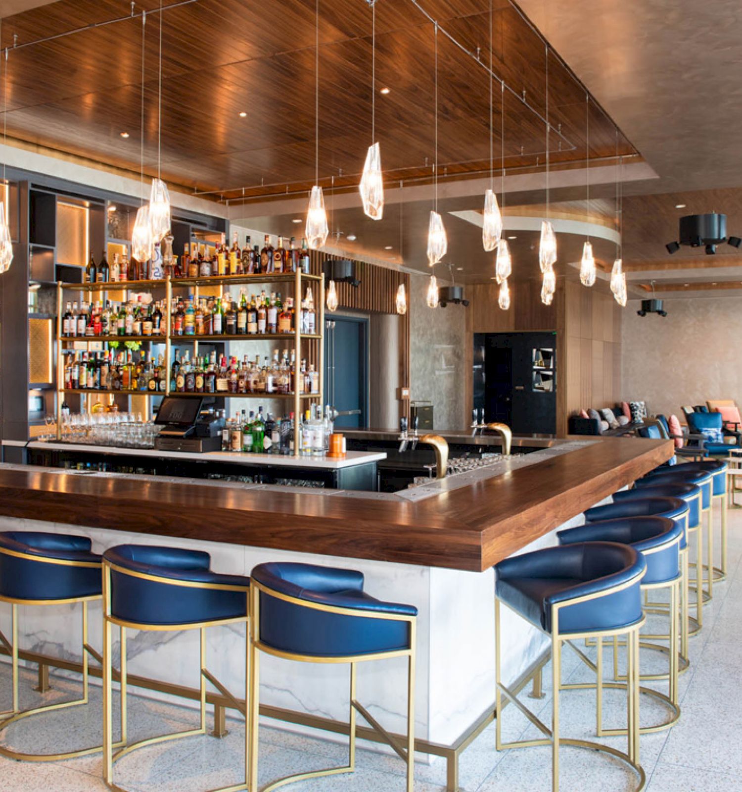 A stylish and modern bar with blue barstools, hanging lights, and a well-stocked shelf of bottles behind the counter, along with a seating area.