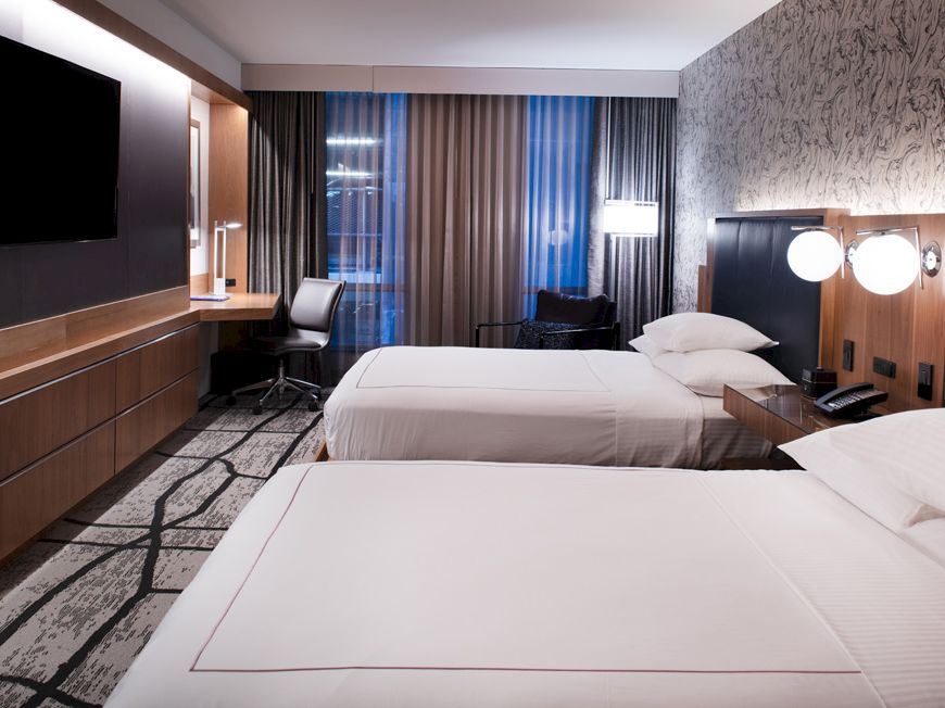 The image shows a modern hotel room with two beds, a wall-mounted TV, a desk, lamps, and large windows with beige curtains.