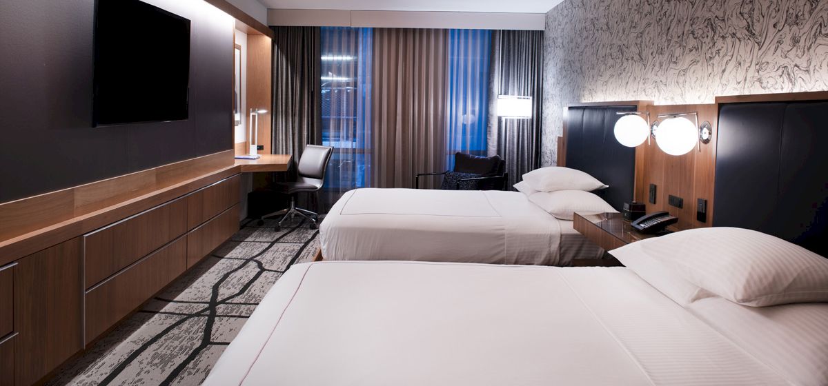 The image shows a modern hotel room with two beds, a wall-mounted TV, a desk, lamps, and large windows with beige curtains.