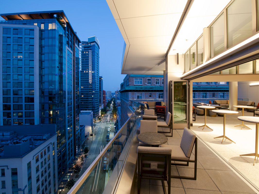 A modern balcony overlooking a city street, with tables and chairs, adjacent to a brightly lit indoor area and tall buildings in the background.
