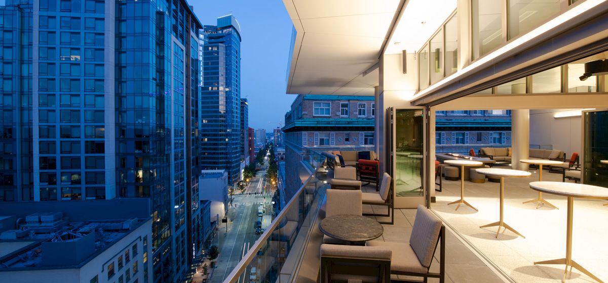 A modern balcony overlooking a city street, with tables and chairs, adjacent to a brightly lit indoor area and tall buildings in the background.