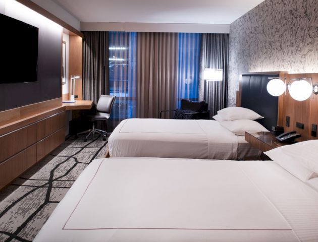 A modern hotel room with two double beds, a flat-screen TV, a work desk, and contemporary decor, featuring large windows and stylish lighting.
