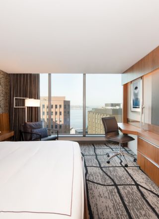 This image shows a modern hotel room with a bed, wall-mounted TV, desk, chair, and a large window offering a city view.