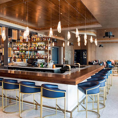 A modern bar with a wooden counter, blue barstools, and hanging lights. The background features a variety of bottles on shelves and seating areas.