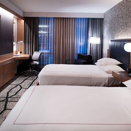 This image shows a modern hotel room with two twin beds, a flat-screen TV, a desk and chair, and large windows with curtains.