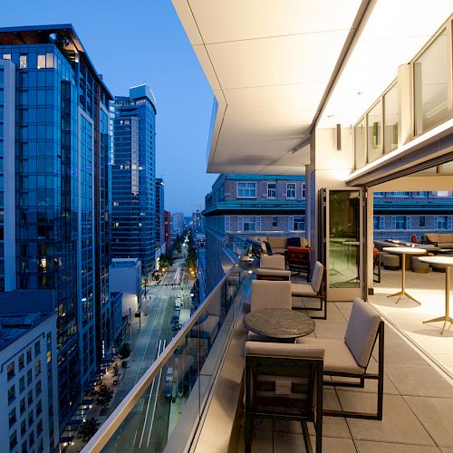 A modern balcony with seating and tables, overlooking a cityscape of tall buildings and a street at dusk, with lights beginning to turn on.