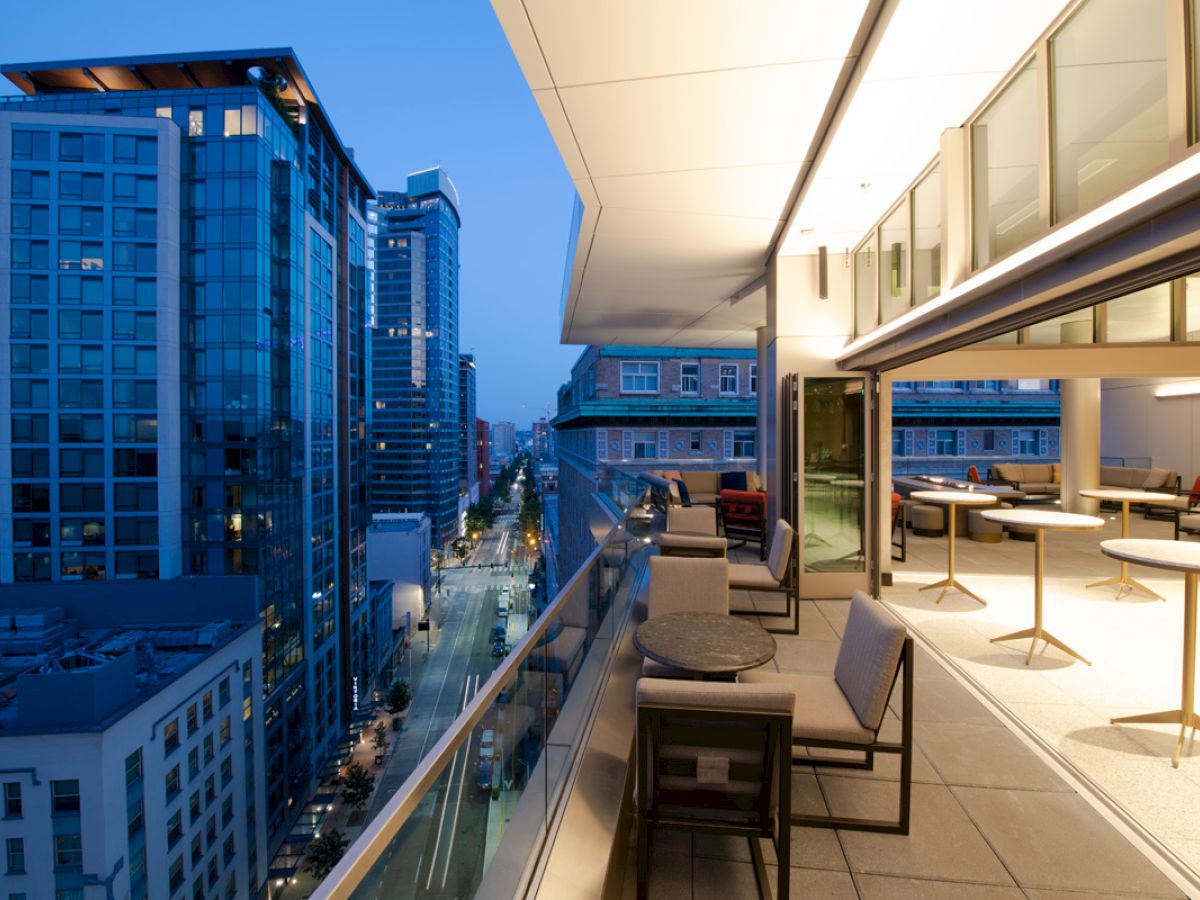 A modern, well-furnished balcony overlooking a street with tall buildings at twilight, featuring seating and tables, with city lights glowing below.