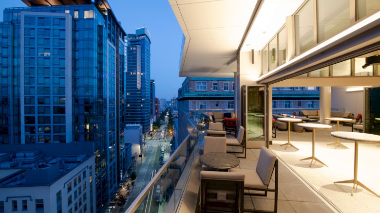 A modern, well-furnished balcony overlooking a street with tall buildings at twilight, featuring seating and tables, with city lights glowing below.