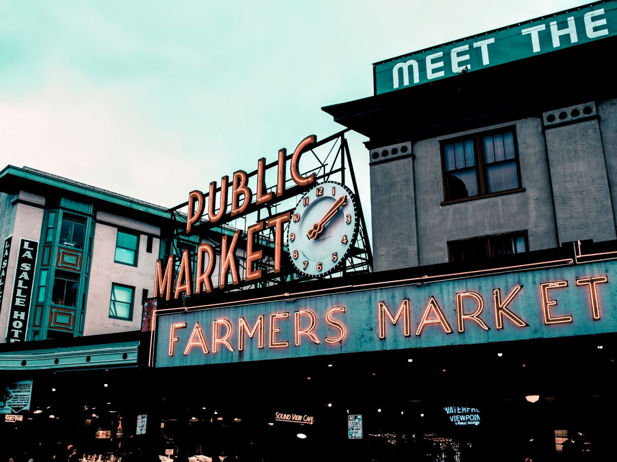 The image shows a neon sign for a public farmers market at Pike Place Market, with a clock and surrounding buildings under a cloudy sky.