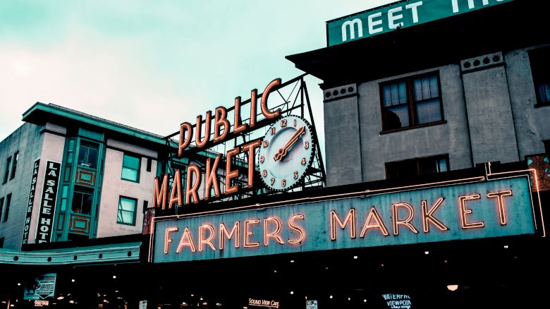 The image shows a neon sign for a public farmers market at Pike Place Market, with a clock and surrounding buildings under a cloudy sky.
