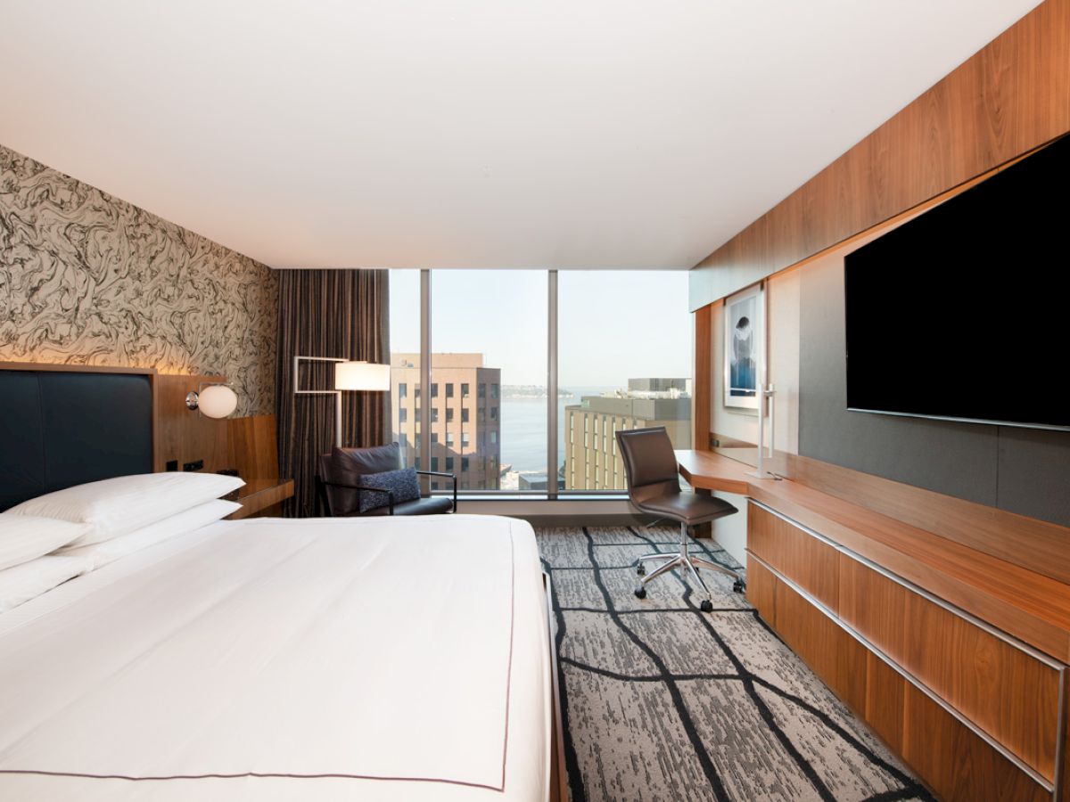 A modern hotel room with a large bed, wall-mounted TV, desk, chair, and a window with a cityscape view in the background.